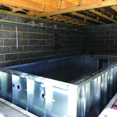 Rectangular galvanized metal pool shell sits recessed on a concrete floor, being installed inside an unfinished basement with exposed wooden joists, cinderblock walls, hanging pipes and a red tool nearby.