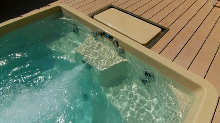 Hot tub corner ejects bubbling jets while sunlight ripples across turquoise water; beige molded rim and control panel with a small unreadable digital display sit flush in light wood decking, metal jet fittings visible.