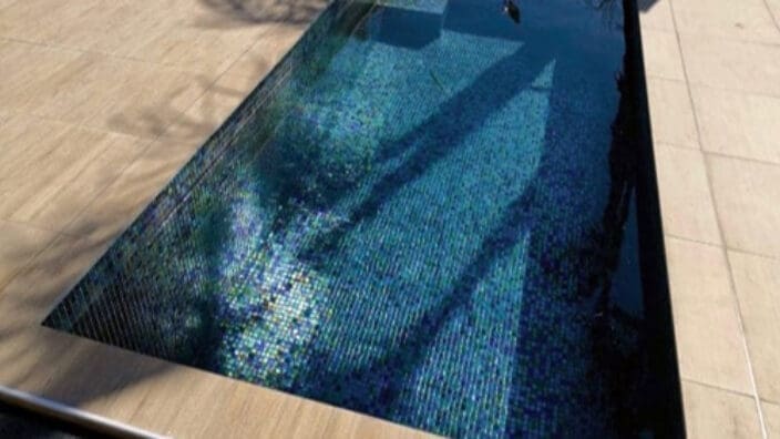 Rectangular tiled plunge pool reflects palm-tree shadows and sunlight, sitting recessed in a light stone patio with stainless edging and surrounding beige paving under a clear daytime sky.