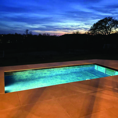 Rectangular in-ground pool glows with turquoise light, reflecting a deep blue twilight sky and silhouetted trees beyond, set within a large tiled patio at dusk.