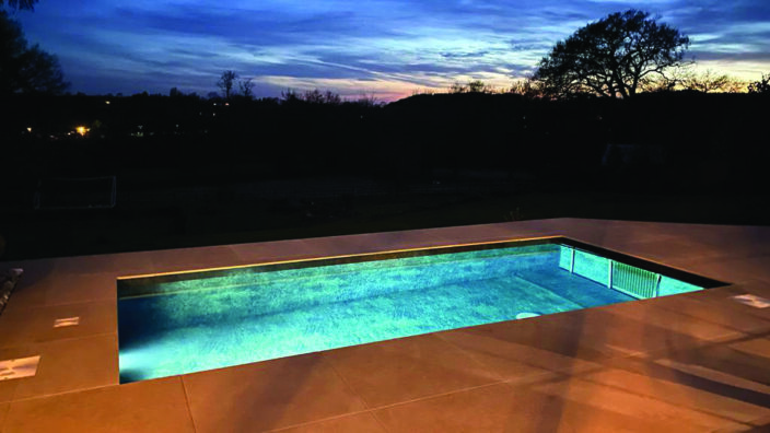 Rectangular in-ground pool glows with turquoise light, reflecting a deep blue twilight sky and silhouetted trees beyond, set within a large tiled patio at dusk.