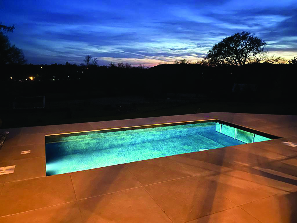 Rectangular in-ground pool glows with turquoise light, reflecting a deep blue twilight sky and silhouetted trees beyond, set within a large tiled patio at dusk.