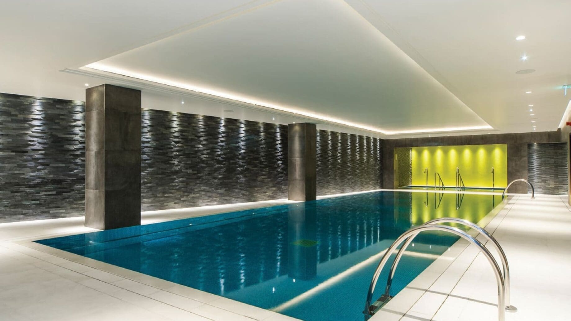 Indoor swimming pool sits still, reflecting recessed ceiling lights; surrounded by white tile deck, dark textured stone walls with pillars, and a yellow-lit alcove at the far end.