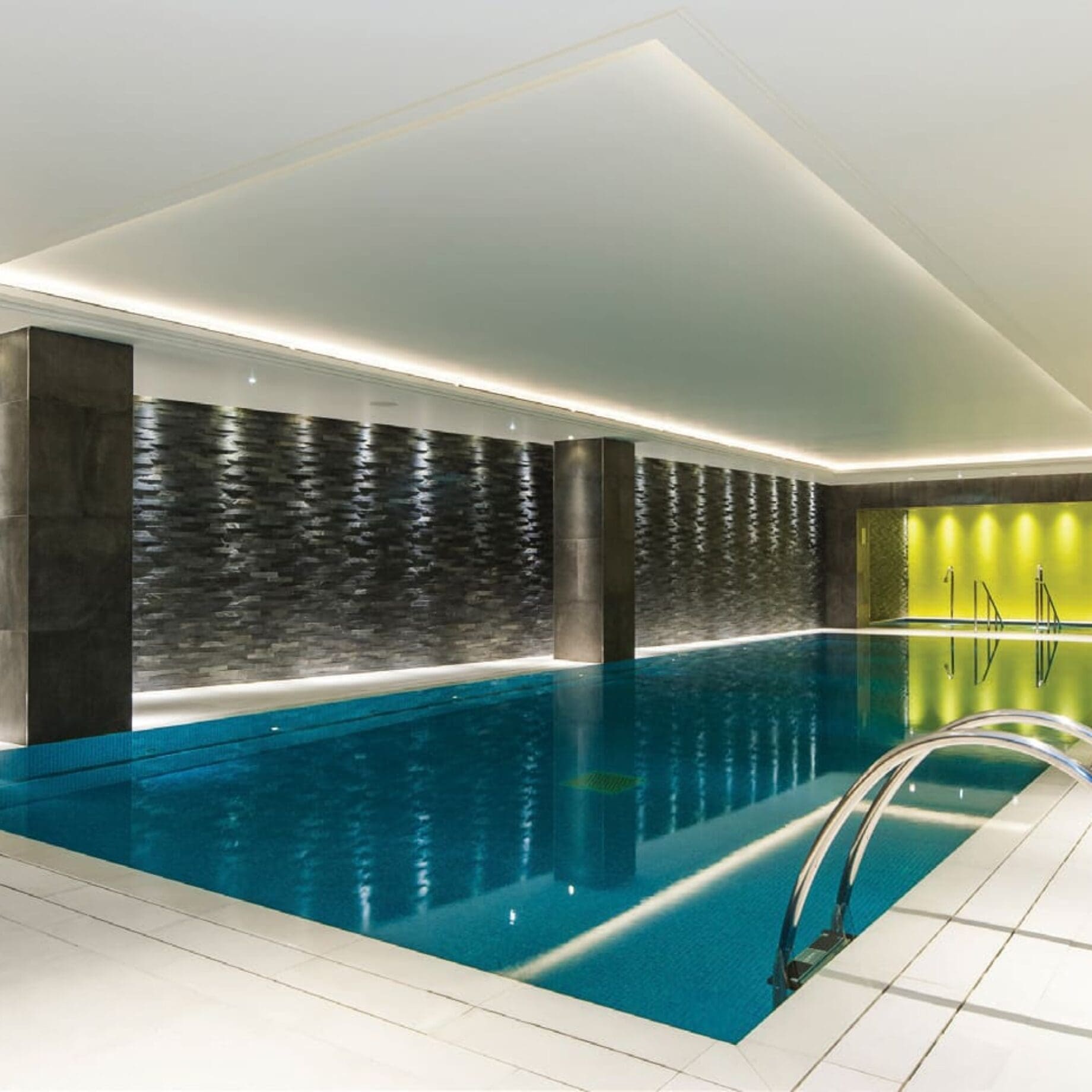 Indoor swimming pool sits still, reflecting recessed ceiling lights; surrounded by white tile deck, dark textured stone walls with pillars, and a yellow-lit alcove at the far end.