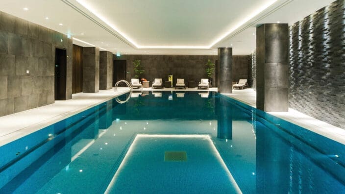 Indoor pool fills the foreground, reflecting ceiling lights; lounge chairs line the far side while tiled pillars and textured walls frame a calm, modern spa environment.