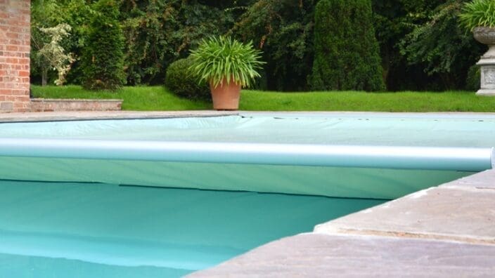 A turquoise pool cover rolls partially across a backyard swimming pool, with stone coping at the edge, brick wall, green lawn, potted plants and an ornamental urn beyond.