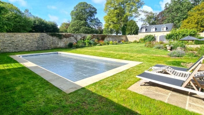 Rectangular covered swimming pool sits in a walled garden, two wooden lounge chairs rest on a stone patio, manicured lawn and flowerbeds surround, stone cottage and tall trees rise beyond.