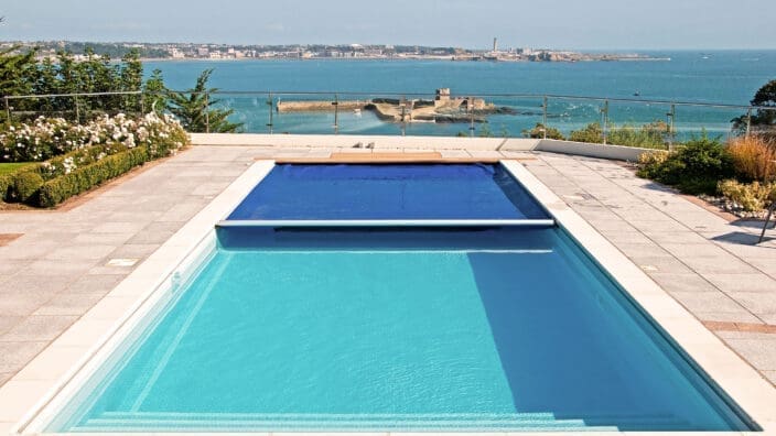 Rectangular pool sits filled with clear turquoise water, partially covered by a blue solar cover, surrounding tiled terrace overlooks a glass-railed coastal vista with a sandy peninsula and distant town.