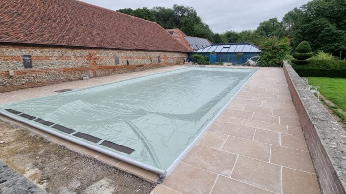 Rectangular outdoor pool covered with a pale-green safety cover, set in a tiled courtyard beside a long flint-and-brick barn, with a manicured lawn, hedges and trees beyond. Text on wall: Shallow End