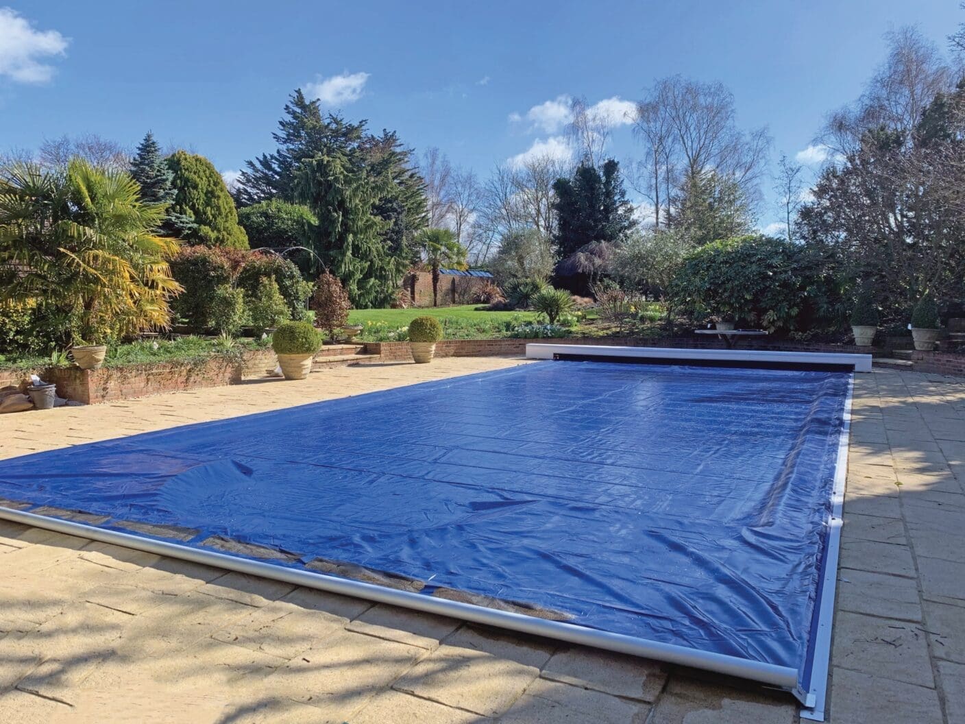 Rectangular outdoor pool covered by a blue solar cover, set on a stone patio with potted shrubs and manicured lawn backed by evergreen and deciduous trees under a clear sky.