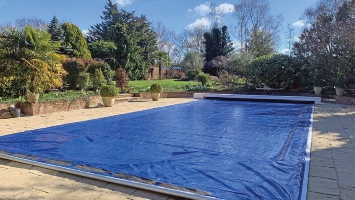 Rectangular outdoor pool covered by a blue solar cover, set on a stone patio with potted shrubs and manicured lawn backed by evergreen and deciduous trees under a clear sky.