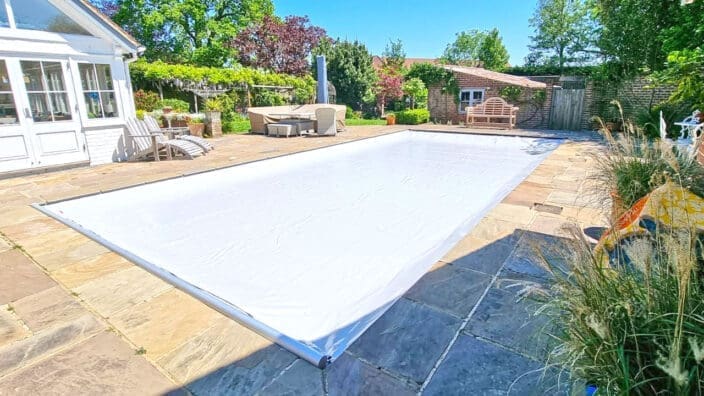 Rectangular pool covered by a taut white cover, set in a stone-paved patio with loungers, wicker seating, potted plants, a white conservatory and brick garden wall under clear sky.