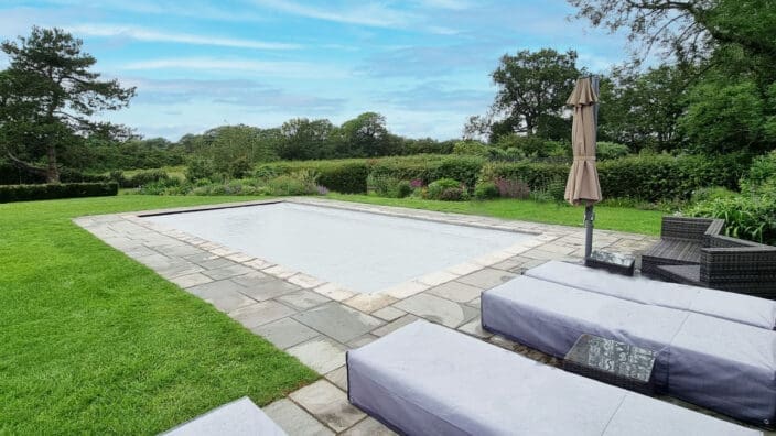 A rectangular covered swimming pool sits unused on a stone patio, flanked by covered lounge beds, wicker chairs and a closed umbrella, surrounded by manicured lawn, hedges and mature trees.