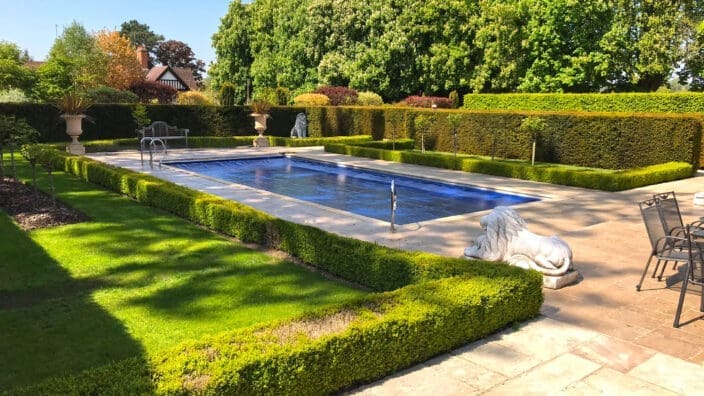 Rectangular swimming pool covered with a blue safety cover sits in a formal sunlit garden, flanked by clipped box hedges, stone lion sculptures, a bench, potted urns and mature trees.