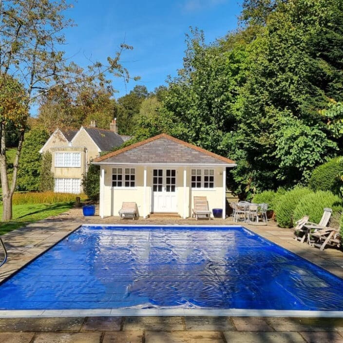 A rectangular outdoor swimming pool covered with a blue cover lies centered on a stone patio, facing a small cream pool house; lawn, sunlit trees and lounge chairs surround it.