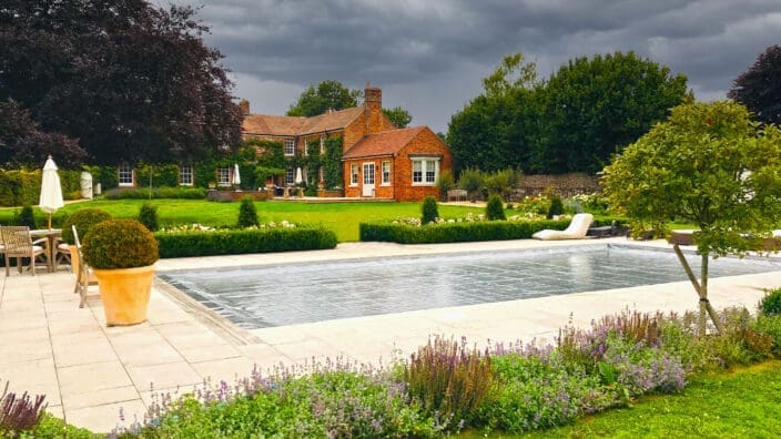 A covered rectangular swimming pool reflects a dark stormy sky, sitting on a paved terrace surrounded by potted shrubs, lounge chairs, flowerbeds and a red-brick country house amid lawns.