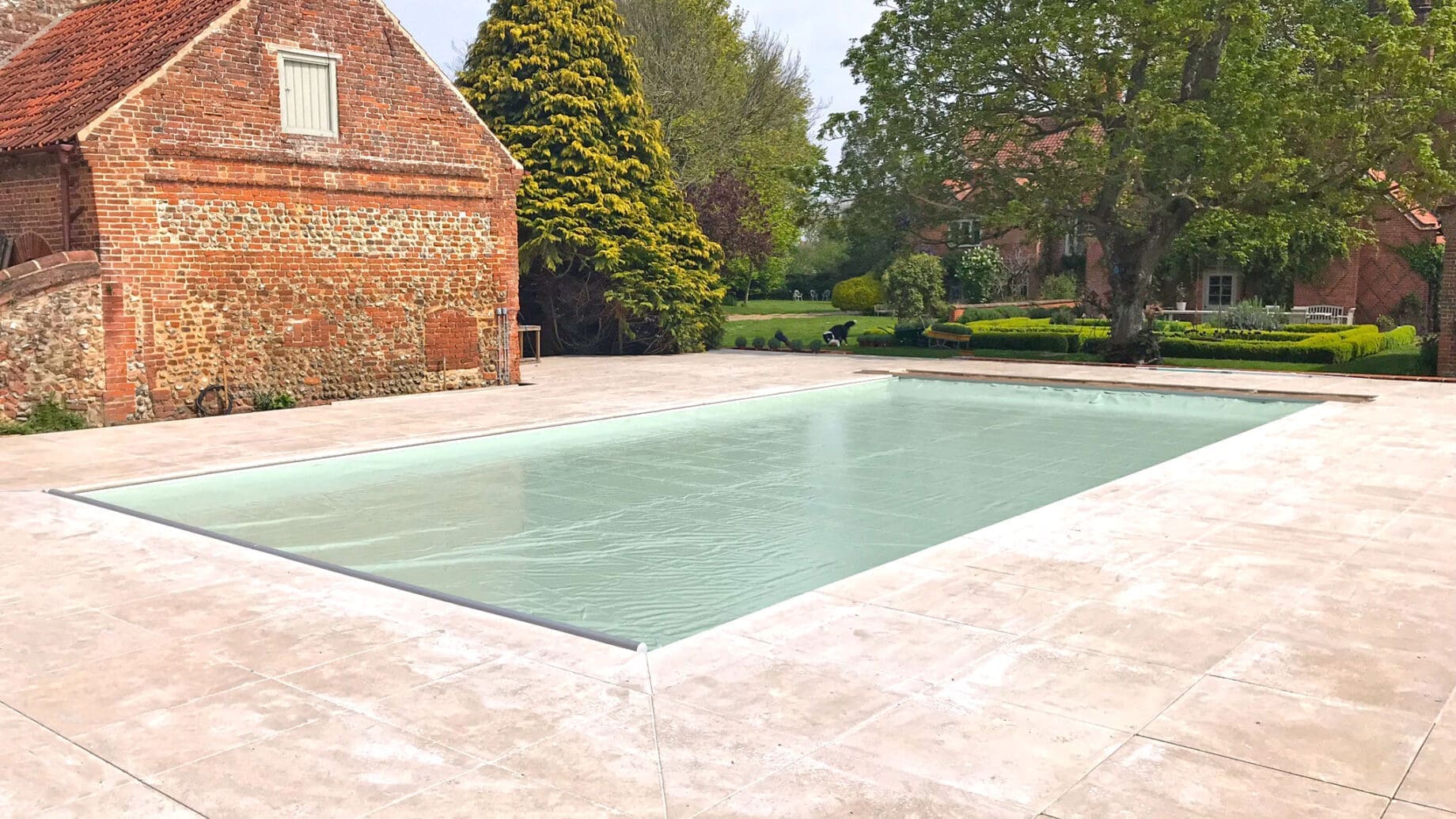Outdoor rectangular swimming pool sits covered with a pale green cover, surrounded by light stone decking, adjacent to an old red-brick barn and a landscaped garden with trees and hedges.
