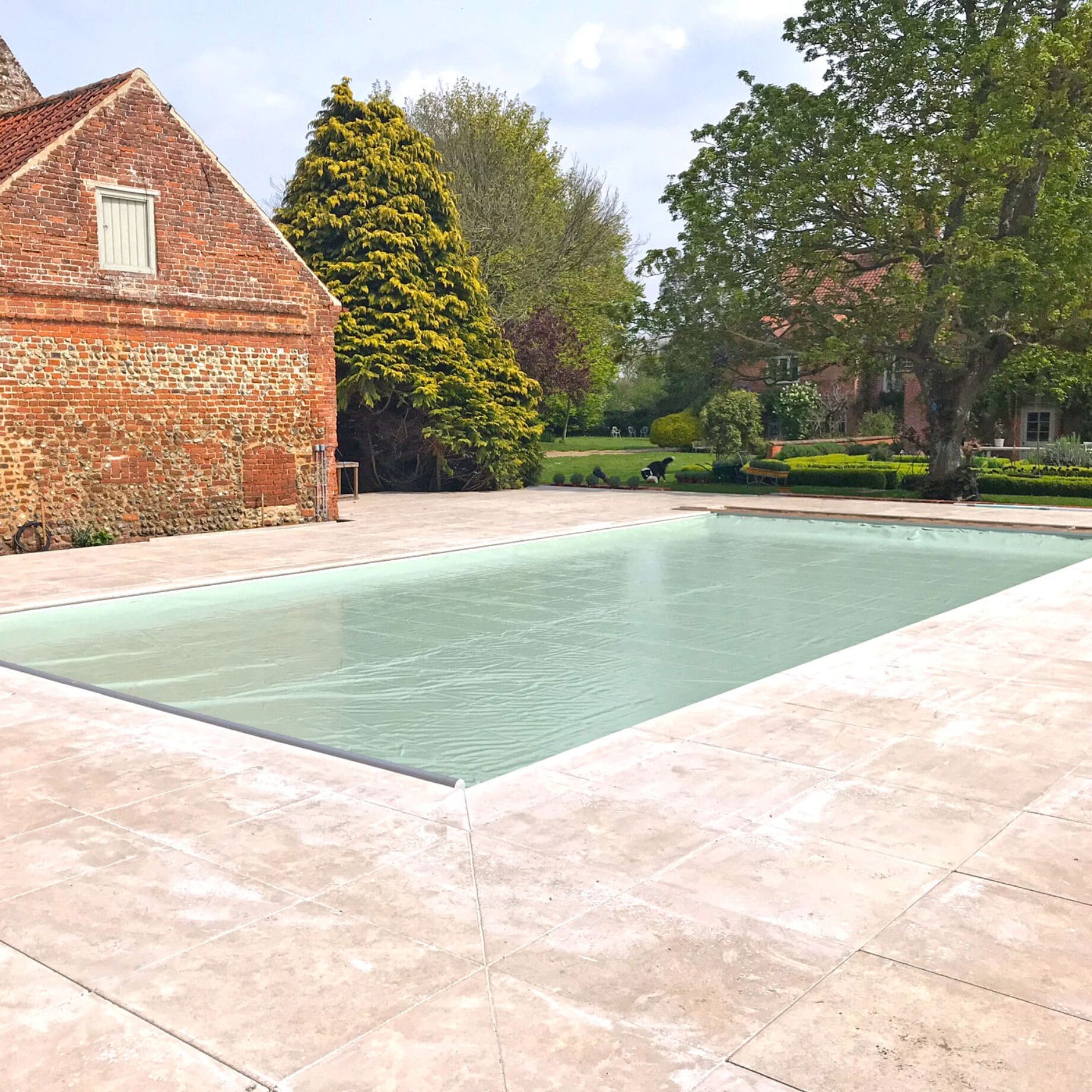 Outdoor rectangular swimming pool sits covered with a pale green cover, surrounded by light stone decking, adjacent to an old red-brick barn and a landscaped garden with trees and hedges.
