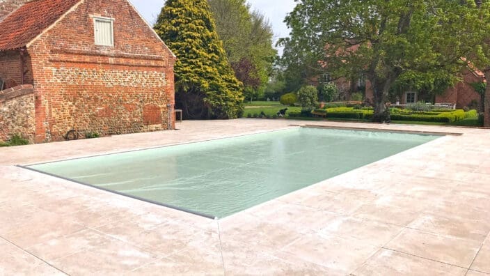 Outdoor rectangular swimming pool sits covered with a pale green cover, surrounded by light stone decking, adjacent to an old red-brick barn and a landscaped garden with trees and hedges.