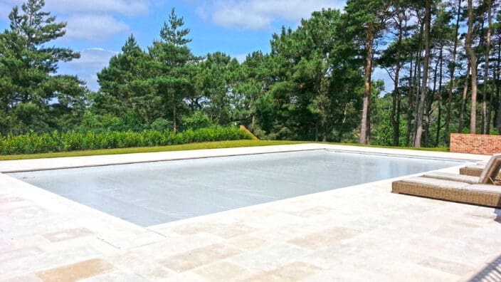A rectangular covered swimming pool sits on a pale stone patio, bordered by wicker lounge chairs, with tall pine trees and blue sky in the wooded background.