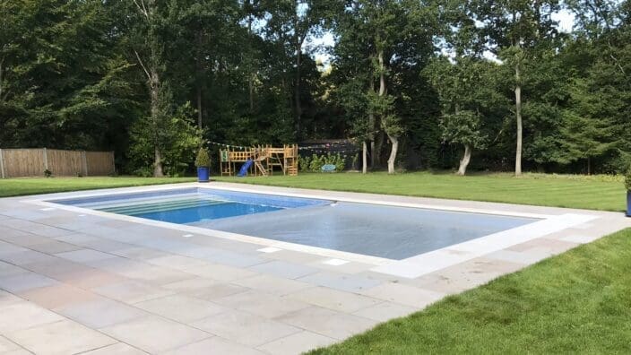 A rectangular in-ground swimming pool sits with calm blue water on a tiled patio, bordered by manicured lawn, a wooden children's playset and dense trees in the backyard.