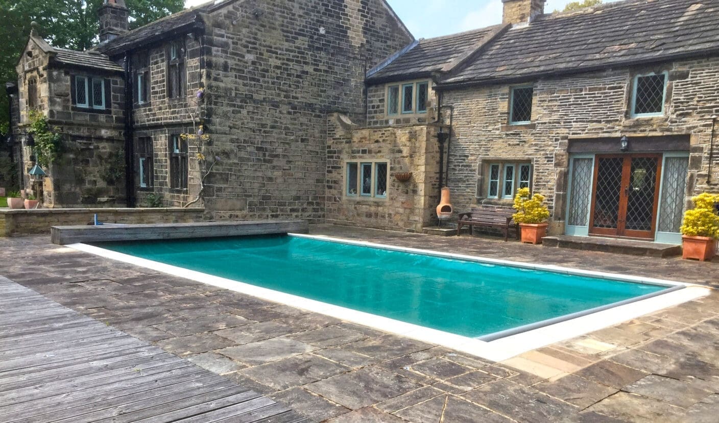 A rectangular swimming pool contains calm turquoise water, bordered by coping and a wide stone patio, adjacent to a stone house with leaded windows, potted shrubs and a wooden bench.