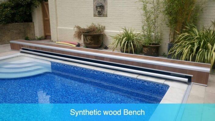 Synthetic wood bench spans pool edge over rolled cover reel; tiled steps descend into blue mosaic pool beside patio with plants. Text in image reads Synthetic wood Bench