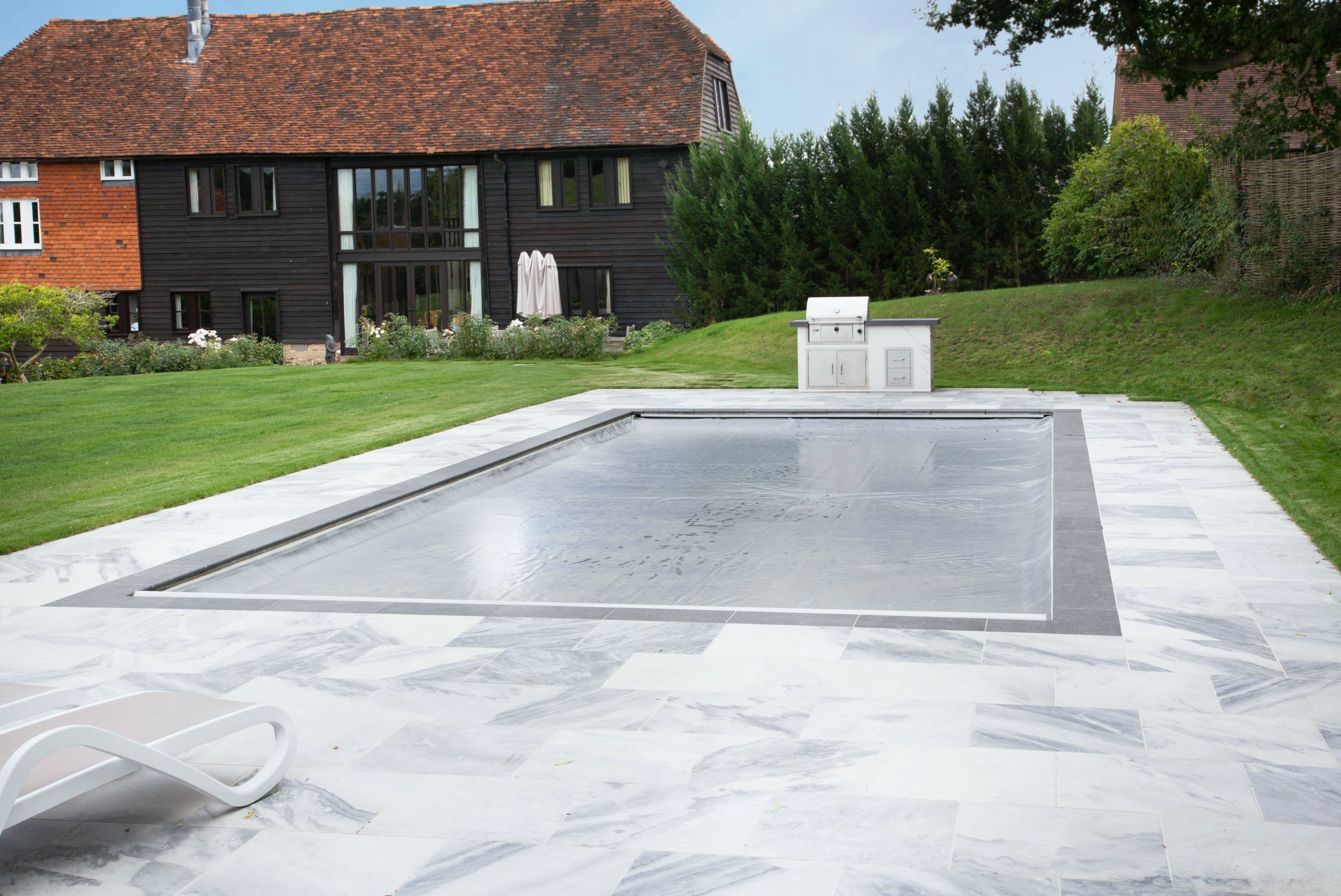 A rectangular pool is covered with a taut safety cover, set within white marble paving beside green lawn, an outdoor grill and a dark timber house with red tiled roof.