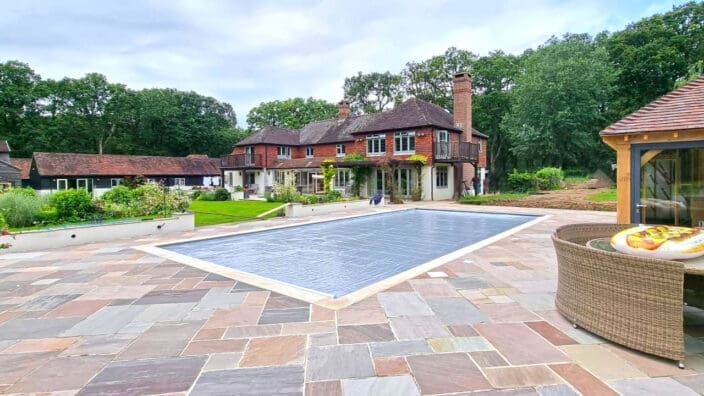 A rectangular covered swimming pool sits on a stone patio, flanked by wicker furniture, with a large brick-and-timber house backing onto a tree-lined lawn.