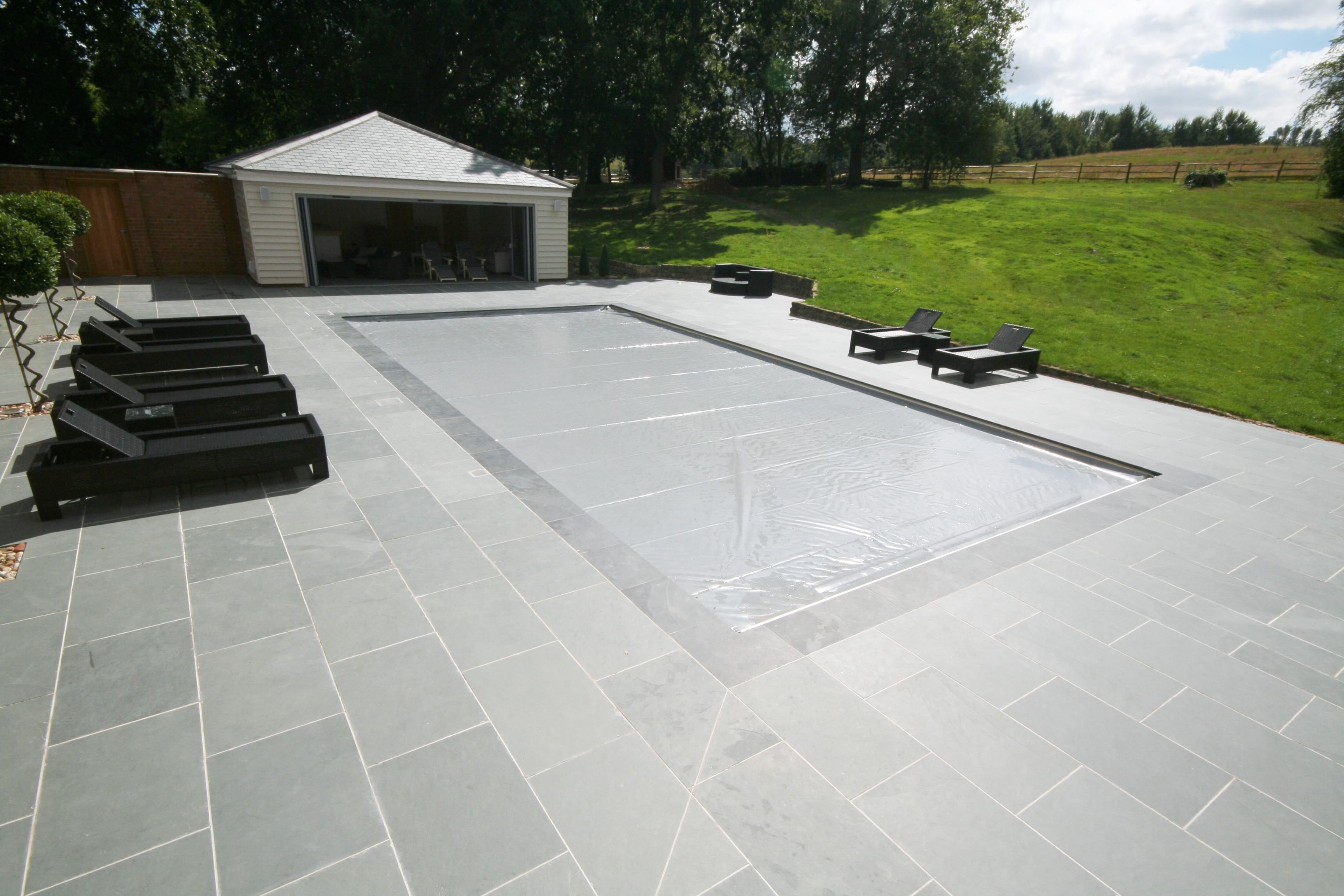 Rectangular in-ground pool covered with a gray protective sheet, surrounded by tiled patio with black lounge chairs, an open pool house at rear and a grassy slope with trees.