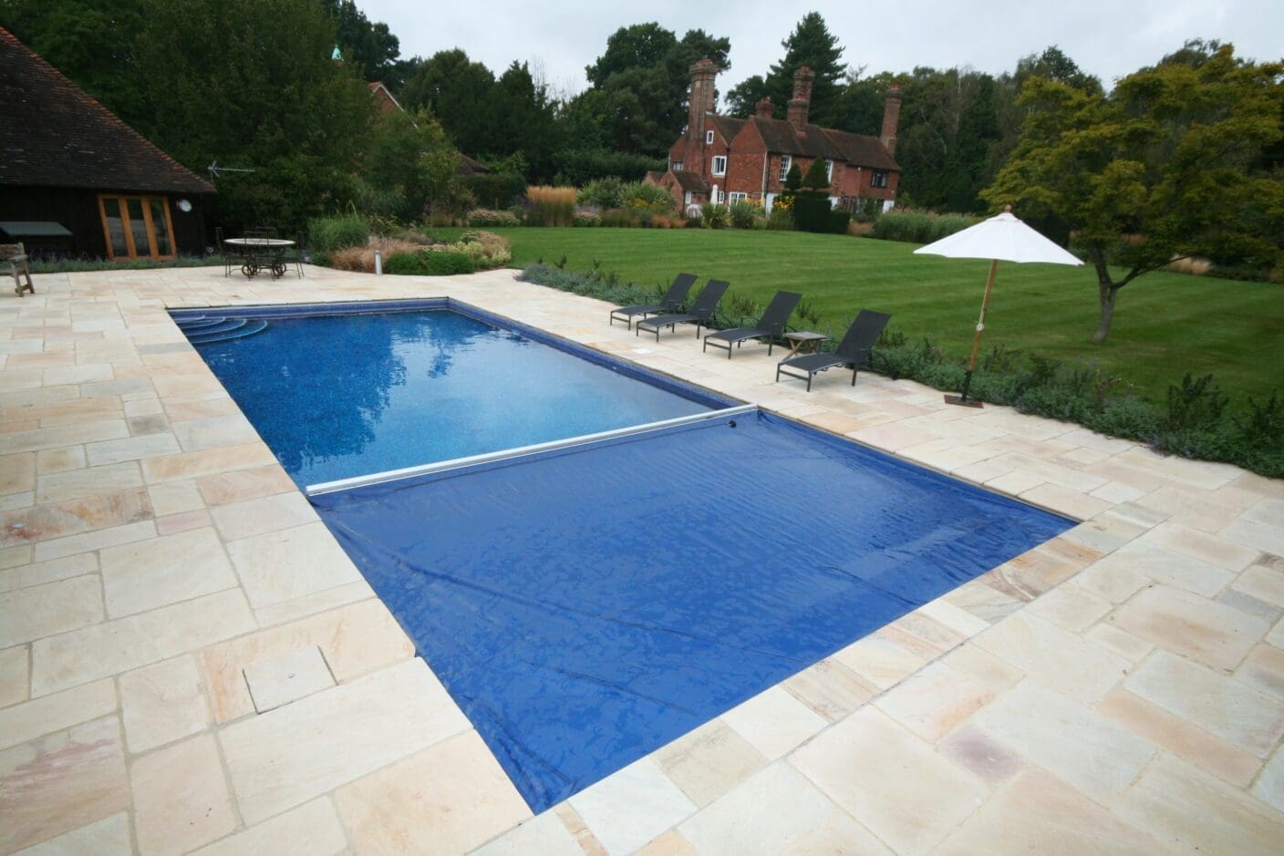 A rectangular swimming pool partially covered by a blue solar cover, set on a pale stone patio with lounge chairs and umbrella beside a large lawn and brick house.
