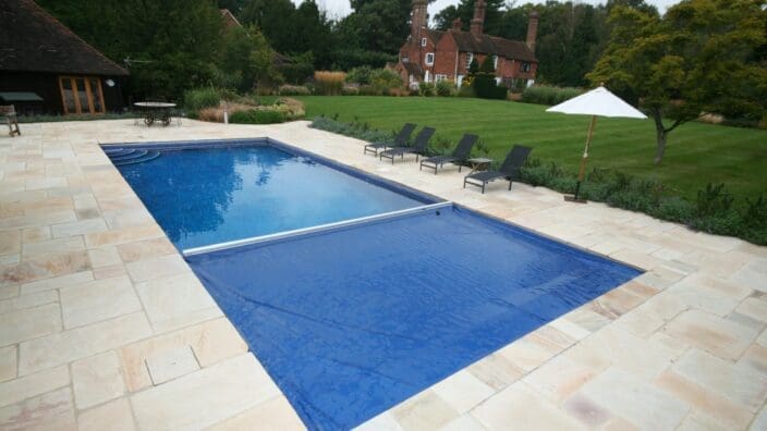 A rectangular swimming pool partially covered by a blue solar cover, set on a pale stone patio with lounge chairs and umbrella beside a large lawn and brick house.