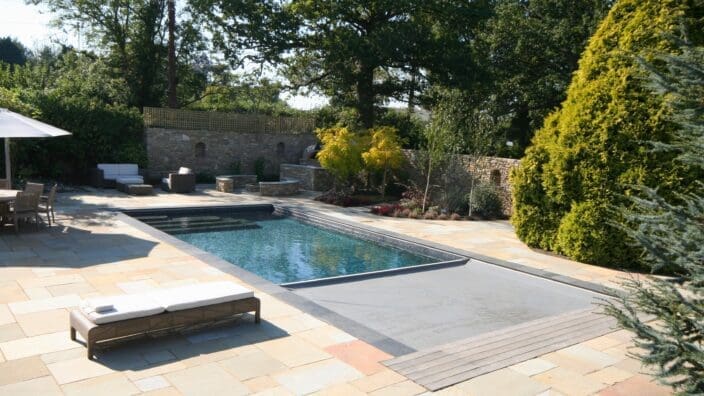 Outdoor rectangular swimming pool sits partially covered by a retractable gray cover while a wicker lounger rests on a stone patio; surrounding trees, shrubs and a stone wall enclose it.