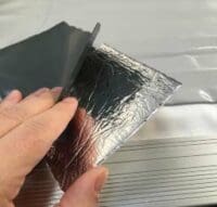 Reflective aluminum foil sheet being peeled back by a hand to reveal an adhesive-backed insulation layer atop a ribbed white surface or panel.