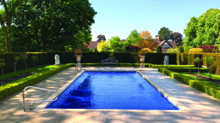 Rectangular swimming pool covered by a taut blue cover, centered on a sunlit stone patio, flanked by low box hedges, urns and lion statues, with trees and a house beyond.