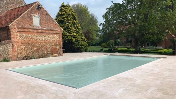 Rectangular outdoor pool covered with a taut pale blue safety cover, set within a wide stone patio, adjacent to an old brick barn and mature trees bordering a manicured lawn.