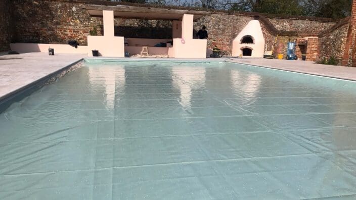 An empty pool is being fitted with a pale turquoise liner, its glossy surface reflecting a pink pavilion and a worker, set within a sunlit stone deck and brick courtyard.