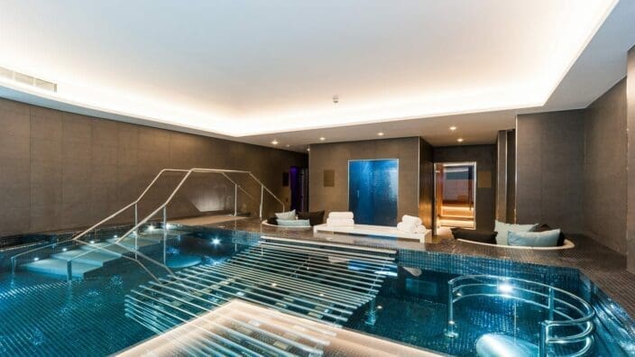 Indoor mosaic-tiled pool with illuminated stainless-steel handrails and submerged loungers, surrounded by cushioned seating, stacked towels, sauna entrance and soft recessed ceiling lighting in a modern spa room.