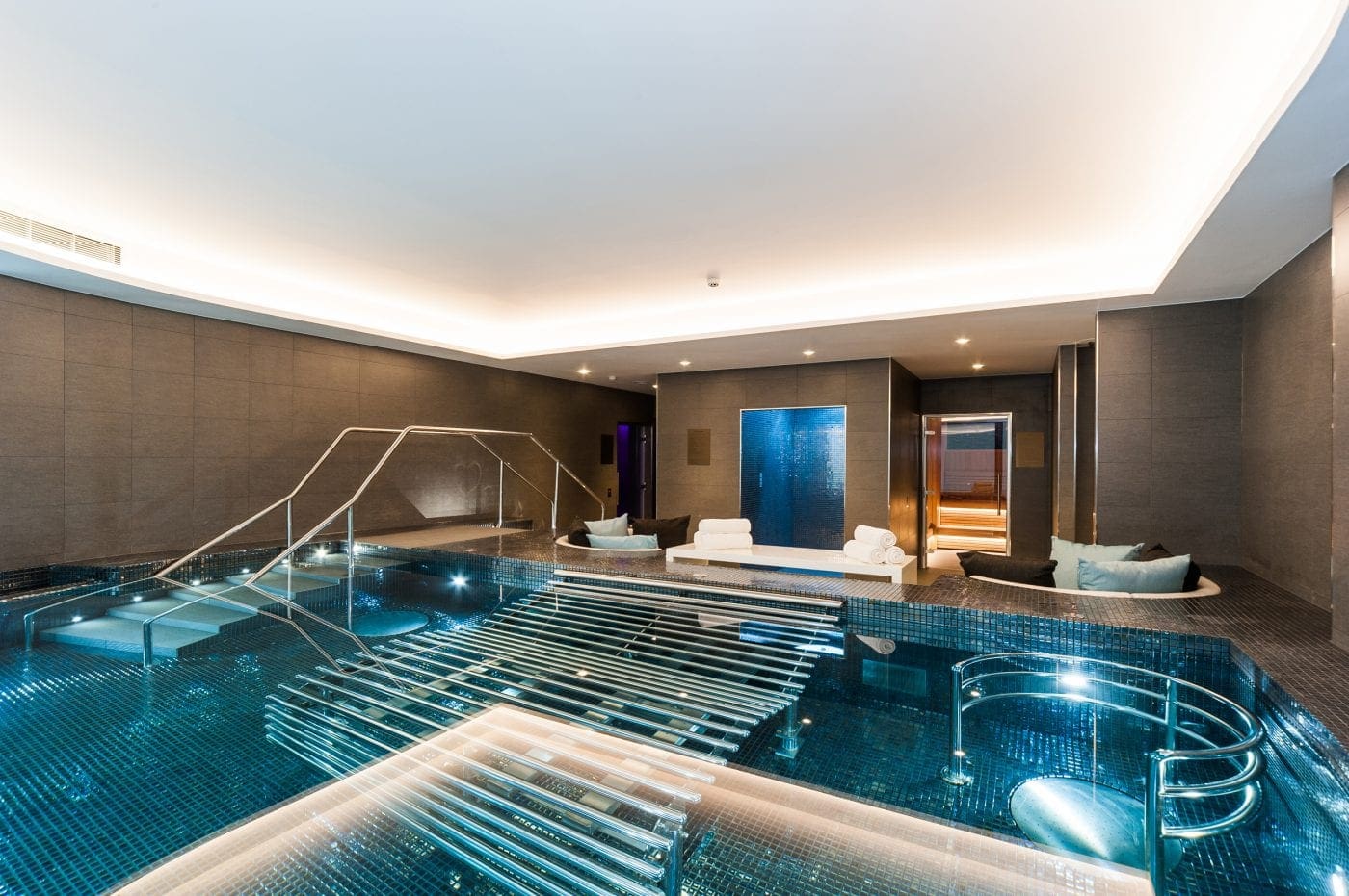 Indoor mosaic-tiled pool with illuminated stainless-steel handrails and submerged loungers, surrounded by cushioned seating, stacked towels, sauna entrance and soft recessed ceiling lighting in a modern spa room.