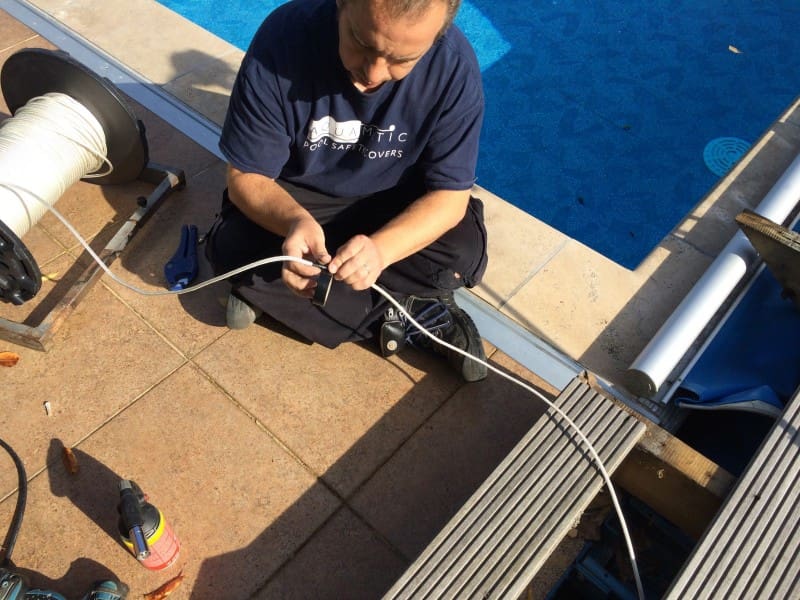 Installer knots a white cord near a pool on a tiled deck, spool and tools nearby; shirt text reads "AQUAMATIC POOL SAFETY COVERS".