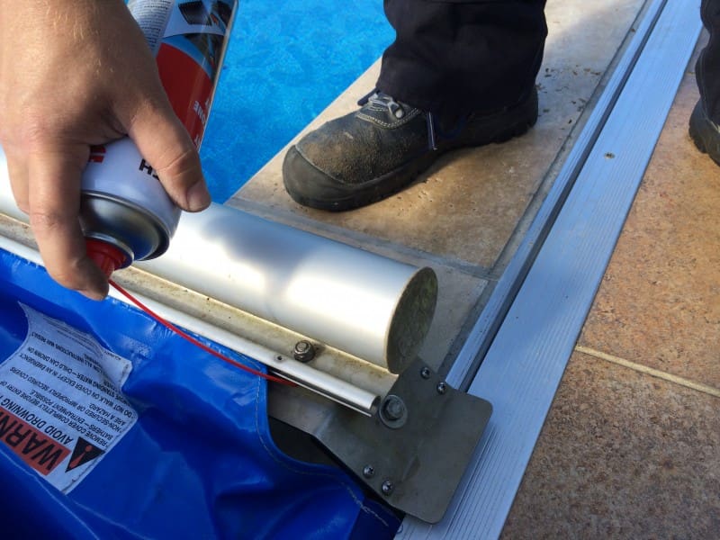 Aluminum pool-cover roller sprayed with aerosol by a hand using a can, positioned on a blue cover at a tiled pool edge with a work boot on the stone decking Text: WARNING AVOID DROWNING