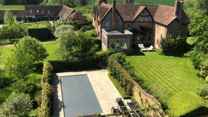 Brick country house sits beside a manicured lawn and a covered rectangular swimming pool, with patio furniture, hedged gardens, trees and neighboring outbuildings set amid rolling green fields.