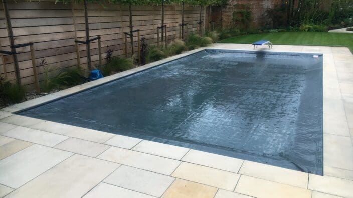 Rectangular inground swimming pool covered by taut dark safety cover, set in pale stone patio with a blue diving board, lawn, young trees and a wooden privacy fence.