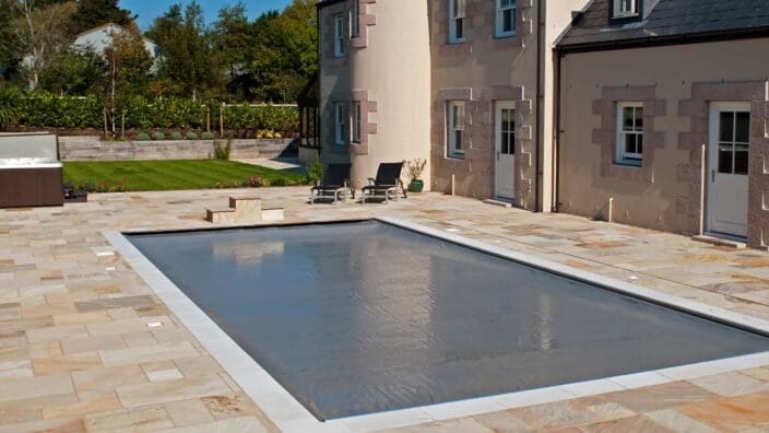 Rectangular in-ground pool sits covered with a taut gray safety cover, surrounded by light stone patio beside a beige two-story house with doors and lounge chairs, lawn and trees beyond.