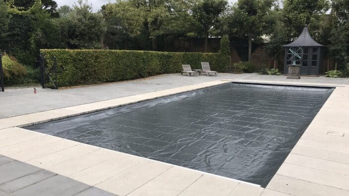 Rectangular outdoor pool sits covered by a dark protective sheet, surrounded by pale stone paving, two loungers, a small gazebo, trimmed hedges and tall trees under a cloudy sky.