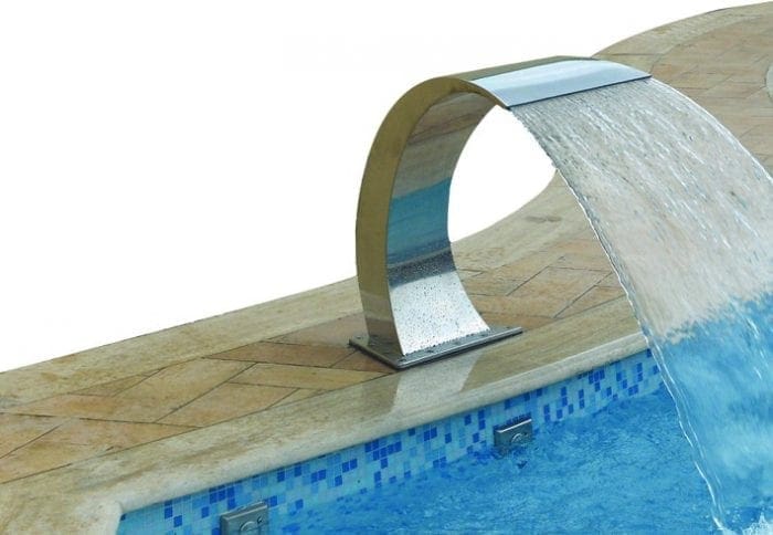 Curved stainless-steel pool fountain arches over the pool edge, pouring a sheet of water into a bright blue mosaic-tiled pool set within a beige stone deck.