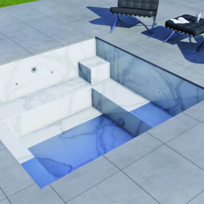 Rectangular marble plunge pool sits recessed in a tiled patio, holding shallow blue water over steps; nearby black leather lounge chairs, hat and drink on the deck, grassy edge beyond.