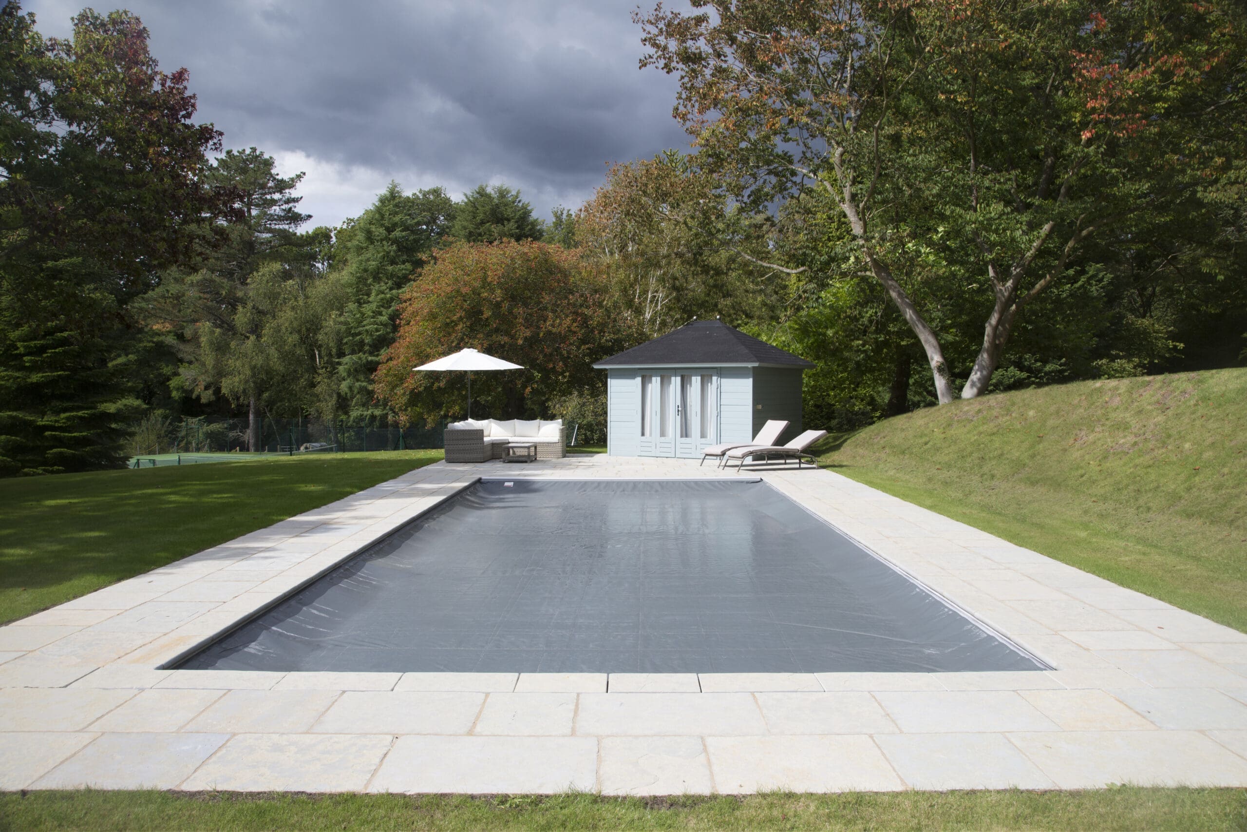 Rectangular pool covered by a tarp sits on a paved terrace with lounge chairs, umbrella, and blue poolhouse, surrounded by lawn and tall trees under a cloudy sky.