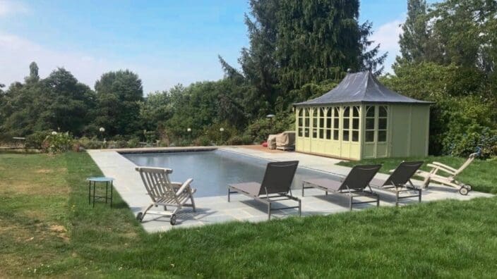 Rectangular swimming pool sits on a stone patio in a grassy backyard, bordered by four gray lounge chairs and two deck chairs, with a pale green pavilion and tall trees beyond.