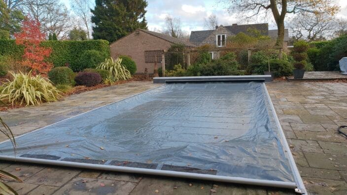 Rectangular swimming pool covered with a translucent plastic pool cover stretched within metal rails, set on a flagstone patio surrounded by trimmed shrubs, potted plants, leaf-strewn ground and houses.